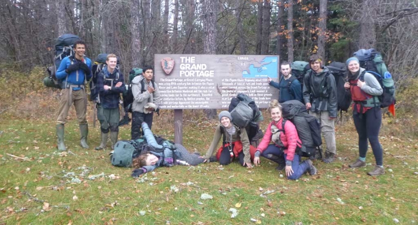 A group of students wearing backpacks pose for a photo with a sign that reads "The Grand Portage," with additional writing, too small to read. 